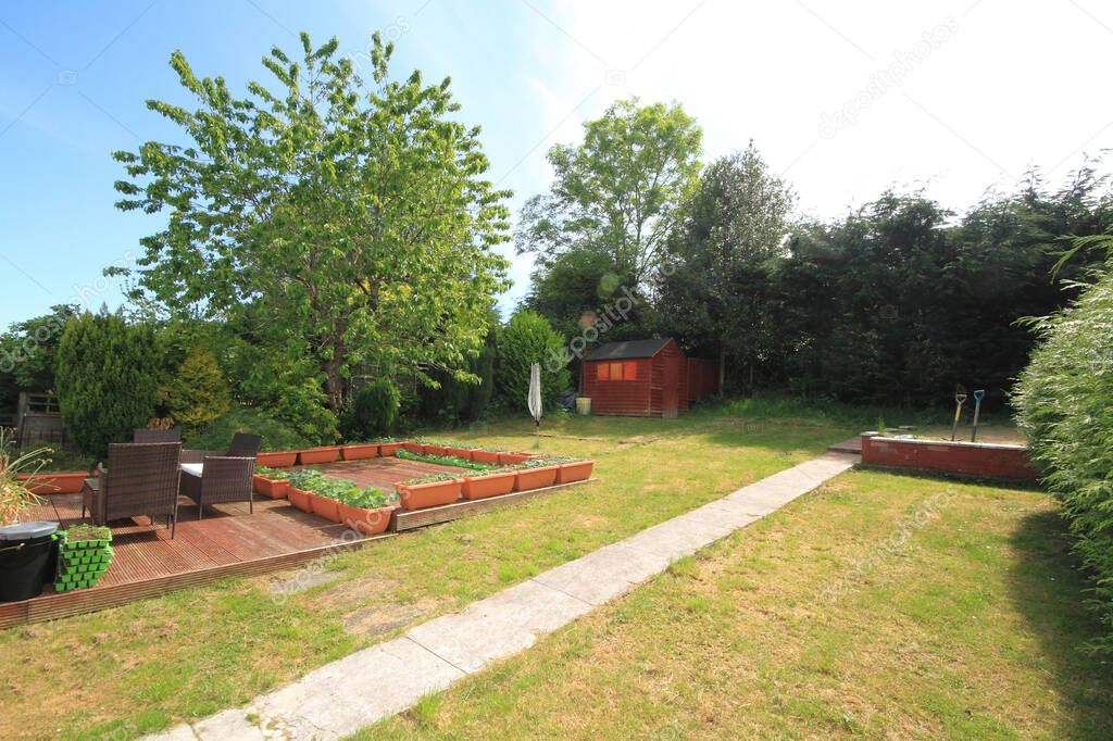 well cared for back garden in summer time with grass, potted flowers, decking area, shed, trees and spades in view. 