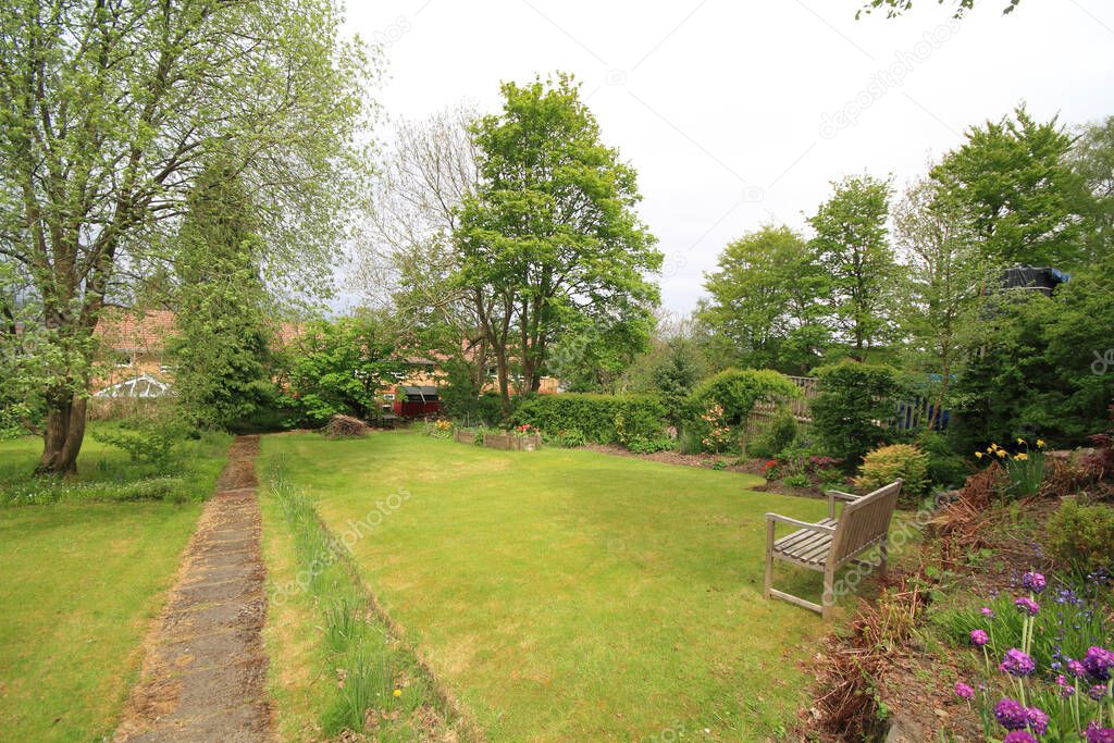 expansive well tended back garden in summer time with bench, trees, flowers and pathway in view.