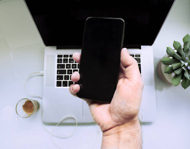 Fotoğraf kesildi, cep telefonunu tutan adamın elinden ve arka planda siyah ekranlı dizüstü bilgisayardan. Boşluğu kopyala..
