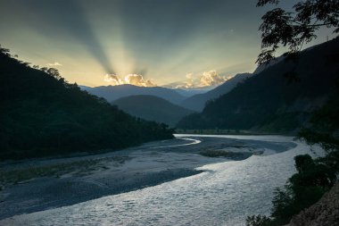 Teesta nehri Himalaya dağlarından akar, güneş ışığı arkaplandaki bulutların arasından gelir, dağ zirvelerinin arkasına güneşi yerleştirir. Sikkim, Hindistan.