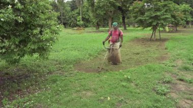 Howrah, Batı Bengal, Hindistan - 20.06.2022 Hintli işçi bahçede çim biçme makinesi kullanarak çim biçiyor. Ağır çekim film.