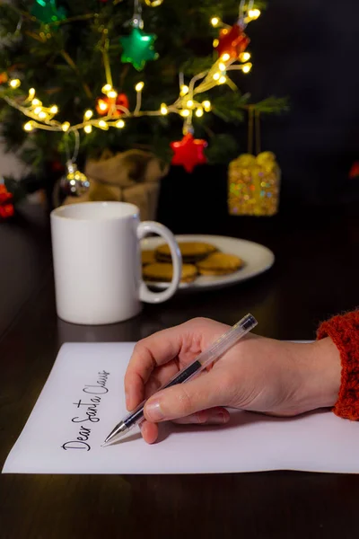 Close-up of a woman's hand writing a letter for Santa Claus. In the background cup of milk, a plate with cookies and a Christmas tree. Christmas and home concept. Vertical photography