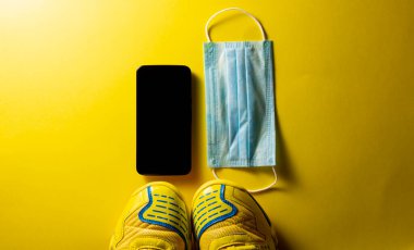 Sarı spor ayakkabılar, akıllı telefon ve cerrahi maske, sarı arka planda. Covid-19 sırasında spor malzemeleri. Boşluğu kopyala