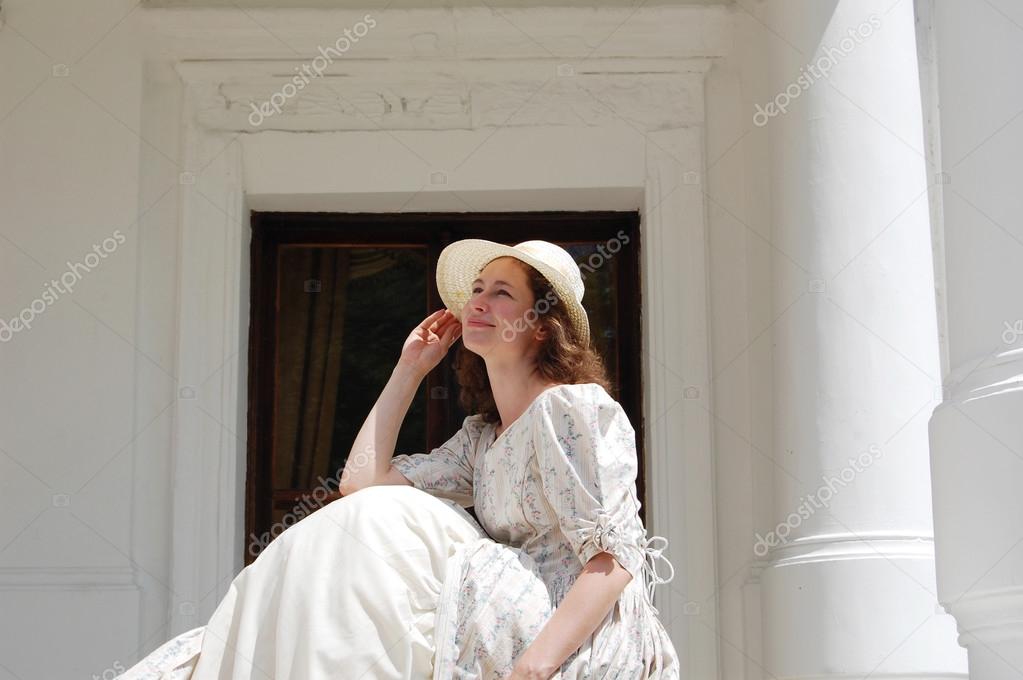 European woman sitting in sunshine and touching hat in vintage dress near palace. 
