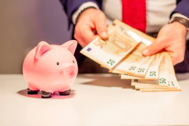 Elindeki elli avroluk banknotla iş adamı, yakın planda domuz kumbarası. Finansal ve para tasarrufu kavramı