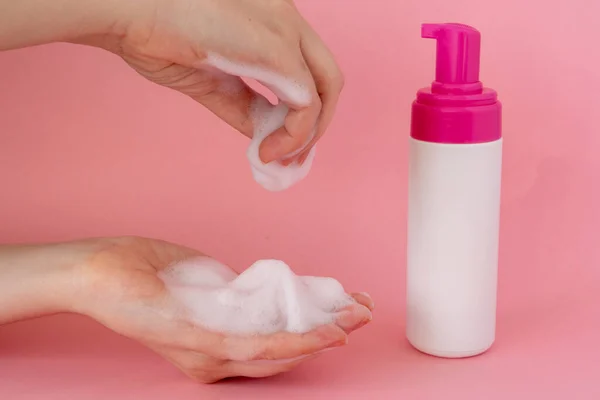 Kadının ellerini yüz köpüğüyle kapatın, pembe arka planda köpüklü köpükle köpük şişeyle.