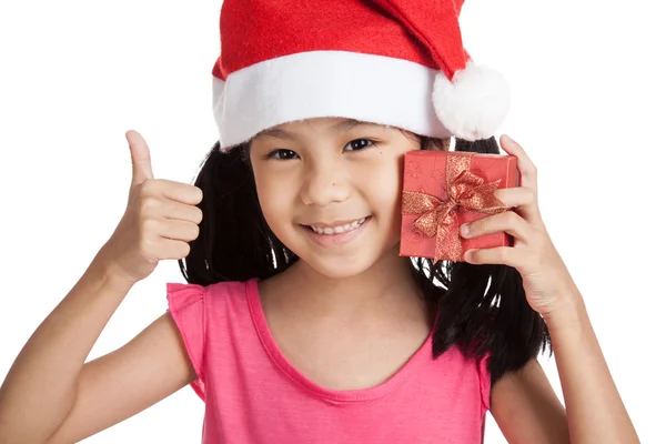 Счастливая маленькая девочка показывает пальцы вверх с шапкой санта и подарком б — стоковое фото