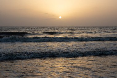 Gökyüzünde turuncu bir parıltı, ufukta güneş batıyor ve okyanusta dalgalar var.