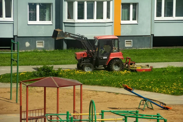 A traktor az udvaron nyírja le a füvet lakóépületek és játszótér mellett. Napos tavaszi nap. — Stock Fotó