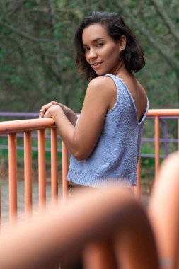 Kısa dalgalı saçlı, güzel bir Latin, korkuluklara yaslanmış, sırtını gösteriyor, mavi bir bluz giyiyor, siyah pantolon giyiyor, gündelik tarzda, parkta.