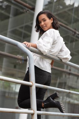 Genç esmer, kısa saçlı, kıvırcık saçlı, sırtüstü uzanmış beyaz bluzu, siyah pantolonu ve botları olan, açık havada