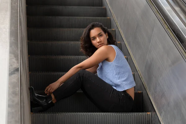 beautiful young latin woman with short wavy hair, sitting on an escalator, wearing a blue blouse, black pants and boots, casual and relaxed style