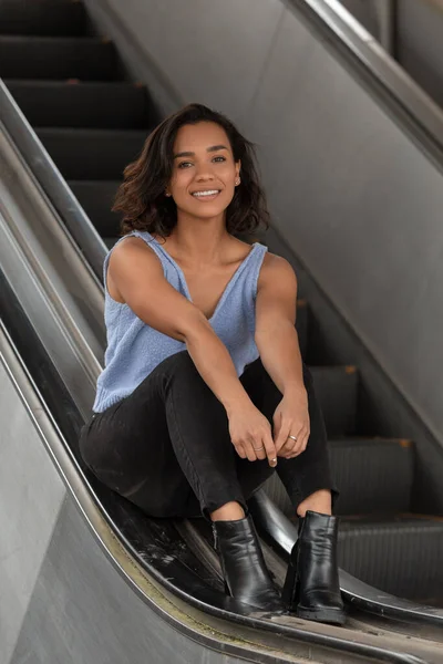 beautiful young latin woman with short wavy hair, sitting on the handrail of some electric stairs, wears a blue blouse, black pants and boots, casual and relaxed style