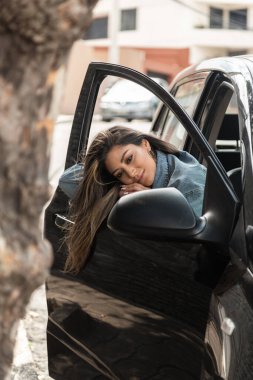 Uzun saçlı ve makyajlı bir Latin kızın yüzü bir arabanın içinde, kollarını cama dayamış, kapı açık ve aynaya bakıyor.