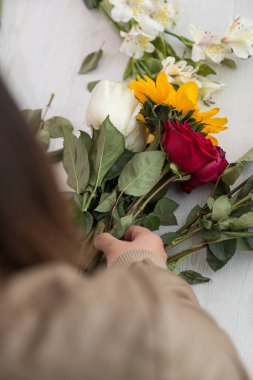 Bir insan elleriyle çiçek aranjmanı yapıyor, ayçiçeği, güller, beyaz bir masanın üzerinde, el işi hediyeler.