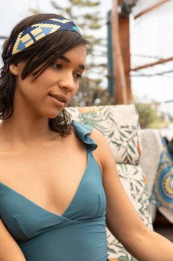 Genç bir kadının yüzü, kısa siyah saçlı, dikkati dağılmış, açık havada duran mavi dekolte bir bluz ve saç bandı giyiyor.