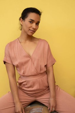 Doğal genç Latin kadın ayakta, pembe günlük bir tulum giyiyor ve saçları toplanmış, sarı arka planı olan bir stüdyo.