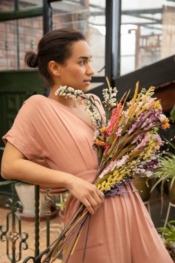 Profilde yüzü olan genç bir manken, renkli bir buket kır çiçeği tutuyor, açık havada günlük kıyafetler giyiyor.