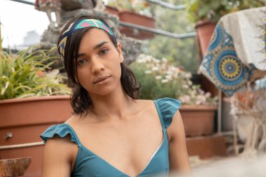 Güzel, kısa saçlı, saç bandı takan, açık havada duran, arka planda bitkiler olan, kısa kesim bir bluz giyen genç bir kadının yüzü.