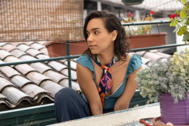 Güzel Latin bir kadın yüzü terasta oturuyor, rengarenk bir eşarp takıyor ve rahat görünüyor.