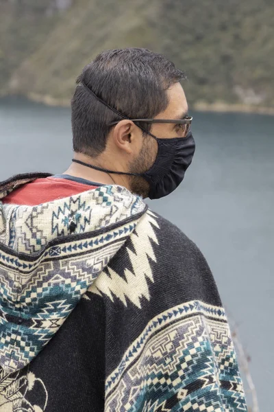 profile of the face of a man with a mask, glasses and a traditional pocho with attractive designs, he is seeing a landscape that has a lake and mountains, nature