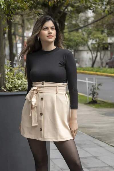 glorious slim, young and latin woman standing with the wind in her long hair, she wears a beige skirt and black blouse, lifestyle and beauty in the background city and trees