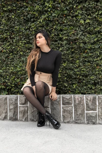 on a sunny day a beautiful young Latin woman with long hair, sitting cross-legged on one edge, wearing casual clothes with a skirt, in the background plants, lifestyle and beauty