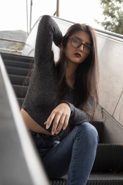 Genç, düz siyah saçlı, gözlüklü, kot pantolonlu ve gri takım elbiseli güzel Latin bir manken, şehirde kolları havada bir şekilde merdivenlerde oturuyor.
