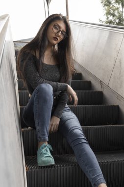 Genç, düz siyah saçlı, gözlüklü, kot pantolonlu ve gri bluzlu güzel Latin bir manken şehirde bir merdivende dinleniyor.