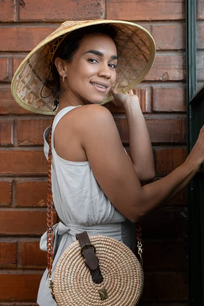 beautiful young brunette woman smiling and posing standing by a brick wall, wears a straw hat and light summer clothes, fashion and lifestyle