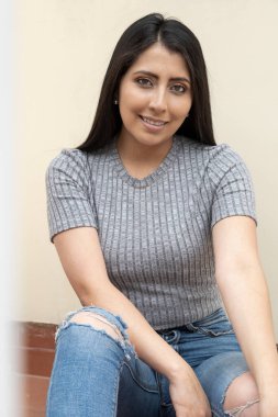 Uzun siyah saçlı, dinlenen, kot pantolon giyen, gri tişörtlü, yüzü doğal makyajlı, yaşam tarzı ve modayla dolu neşeli genç bir manken.