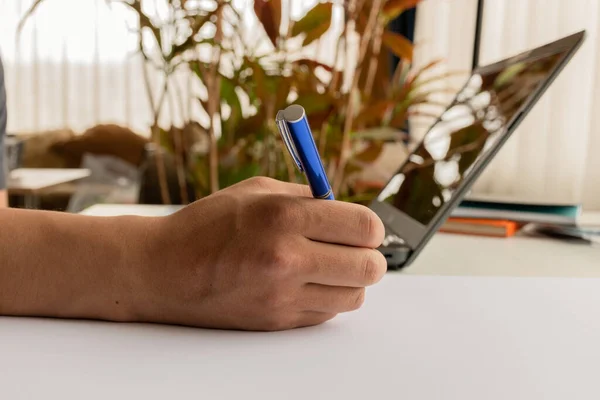 modern laptop next to a hand that is holding a pen to write on a desk, detail of objects and work, business lifestyle