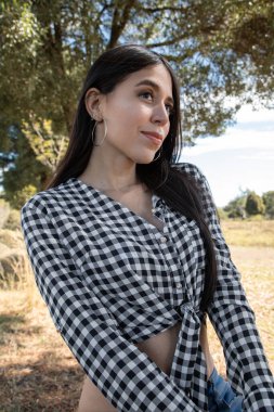 Uzun siyah saçlı, güzel bir kadının park yüzünde, ekose bir bluz giyiyor, arka plandaki ağaçlarda, güzellik ve doğa dinlendirici bir günde.