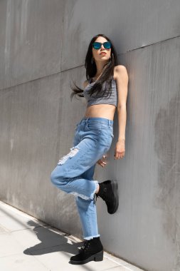 Genç, ince ve güzel bir kadının vücudu güneşli bir yaz gününde duvara yaslanıyor, kot pantolon, üst ve güneş gözlüğü giyiyor, gündelik ve modern tarzda, güzellik dolu bir yaşam tarzı.