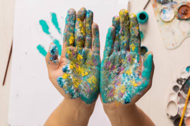 Bir sanatçının çalışma alanının ayrıntıları, paletler, fırçalar, kağıt, boya, sanat ve yaratıcılık, yaşam tarzı gibi çeşitli çalışma araçlarıyla boyanmış avuçlarını gösteren bir masa.