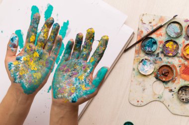 Paletler, fırçalar, kağıt, boya, sanat ve yaratıcılık, stüdyo yaşam tarzı gibi çeşitli çalışma araçlarıyla dolu bir masanın üzerinde ellerini boyayla boyayarak çalışan sanatçı