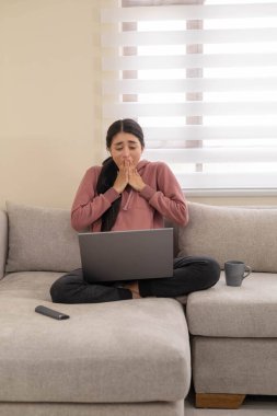 Genç bir kadın dizüstü bilgisayarını kanepede kullanırken hayal kırıklığı içinde bağırıyor. Dijital stres, tükenme ve teknolojiye duygusal tepki kavramı.