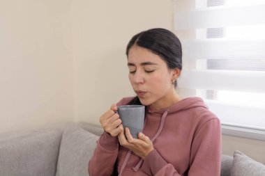 Genç bir kadın rahat bir koltukta oturuyor, elinde sıcak bir fincan kahve tutuyor ve evde sakin bir sabahın tadını çıkarıyor..