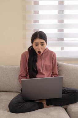 Kanepede bacak bacak üstüne atmış bir kadın dizüstü bilgisayarına öfkeyle tepki veriyor. Stres sembolü, dijital yorgunluk veya teknoloji hüsranı..