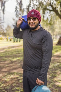 Gülümseyen adam her iki kolunda bir uyku tulumu tutuyor, bir orman kampında maceraya hazır bir şekilde yürüyor..