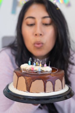 Gülümseyen kadın evde doğum gününü kutluyor, çikolatalı pastasının üzerindeki mumu üflüyor. Etrafı abur cubur ve hediyelerle çevrili..