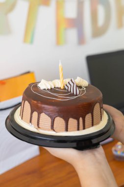 Çikolatalı doğum günü pastasının yakın çekimi, üzerinde krema ve bir mum var, etrafı keklerle çevrili ve ahşap bir yüzey üzerinde hediyeler, samimi bir kutlamayı sembolize ediyor..