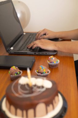 Çikolatalı pasta, kremalı ve mumlu. Yanında renkli kekler ve ahşap bir masada hediye paketleri var. Ev ofis kutlaması için hazır..