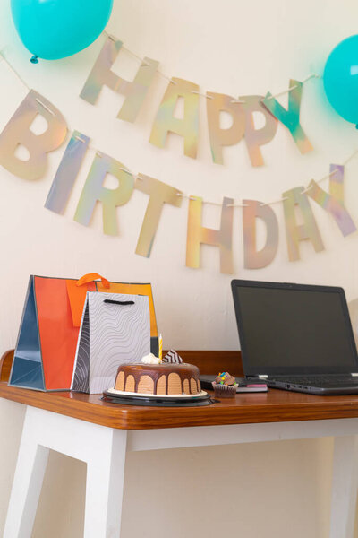 Яркая и современная домашняя сцена с деревянным столом с ноутбуком, шоколадным тортом, подарочными пакетами и воздушными шарами под баннером "Happy Birthday". Минималистичная и радостная настройка для цифрового дня рождения или удаленного празднования.
