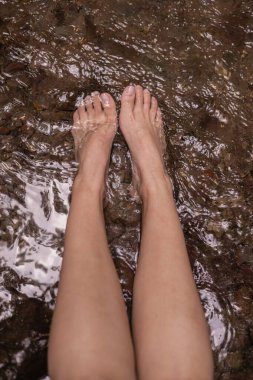 Nehir kıyısında ayaklarını rutubetli taşlara koyup su ve doğa ile temasın doğal ve huzurlu anını yakalayan bir kadının yakın plan çekimi..