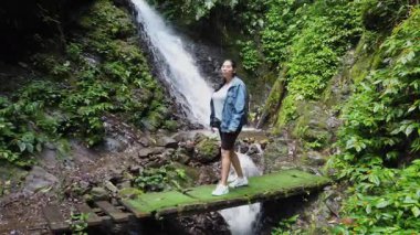 Yeşil bitki örtüsüyle çevrili yosunlu ahşap bir köprüde duran bir kadın ormandaki güçlü bir şelaleyi gözlemliyor..