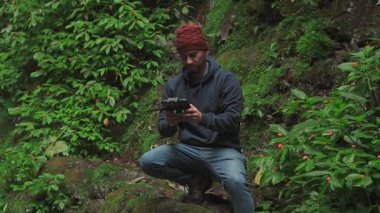 Bir adam, canlı ve nemli bir ormanı keşfederken kamerasını kontrol etmek için bir kayanın üzerinde duruyor..