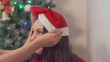 Genç bir kadın, Noel Baba şapkasını dekore edilmiş bir Noel ağacının önünde arkadaşına ayarlıyor, tatlı ve neşeli bir tatil sahnesi yakalıyor..