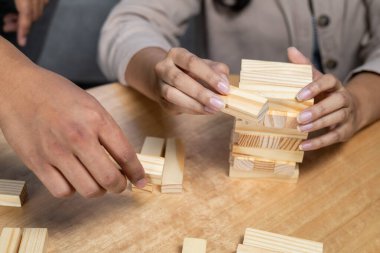 Bir kadın ve erkeğin dikkatlice analiz ettiği ve kırılgan bir istif kulesinden ahşap bir blok seçtiği, odak, strateji, denge ve gerilim vurgulandığı kapalı bir masa üstü oyunu sırasında.