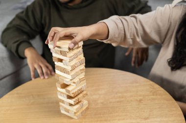 Bir kadının eli dikkatle denge, hassasiyet ve kontrol üzerine odaklanmış gergin bir masa başı oyunu sırasında kırılgan bir istifleme kulesinin tepesine tahta bir bloğu yerleştirirken..